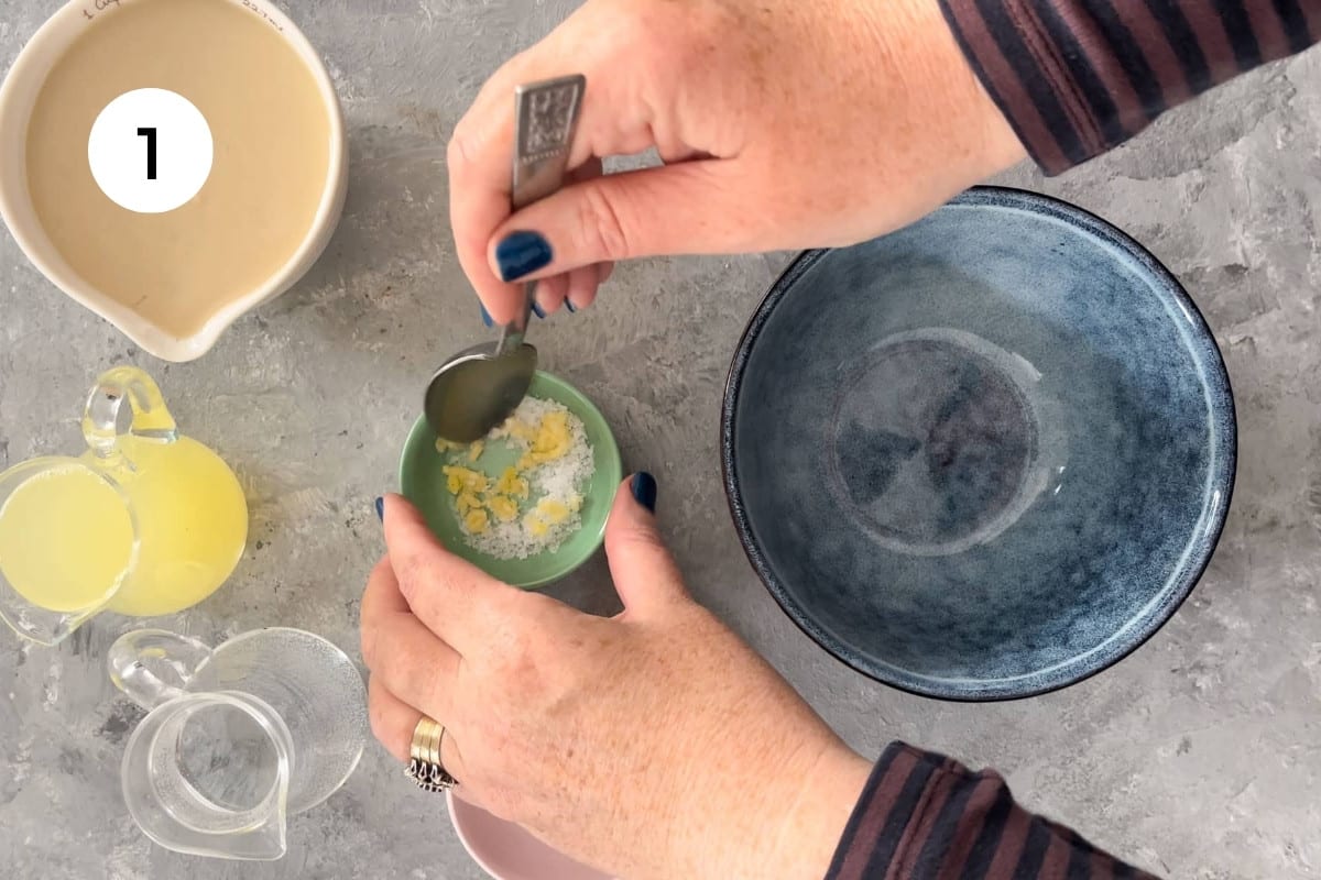 A white hand is stirring crushed garlic and salt together in a small green bowl with a silver spoon.