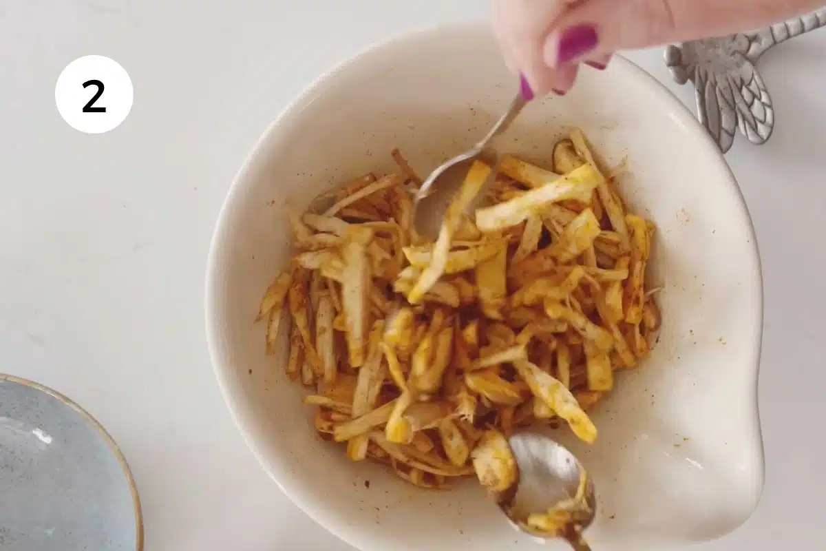 Two white hands are using silver spoons to mix oil and spices into the shredded mushrooms.
