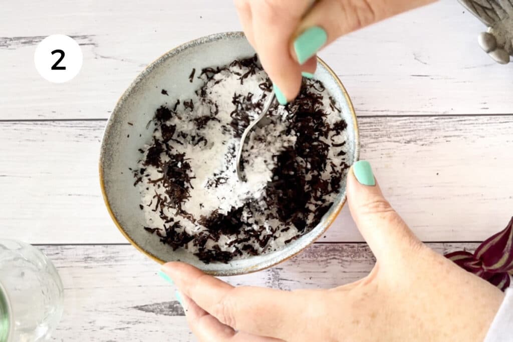 A white hand is using a silver spoon to mix grated truffle and sea salt together in a bowl.