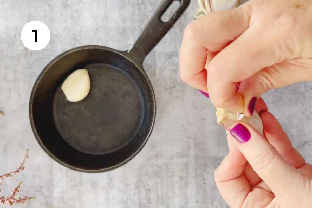 Two white hands with pink/purple nail polish are peeling a clove of garlic. A small cast iron frying pan is close by.