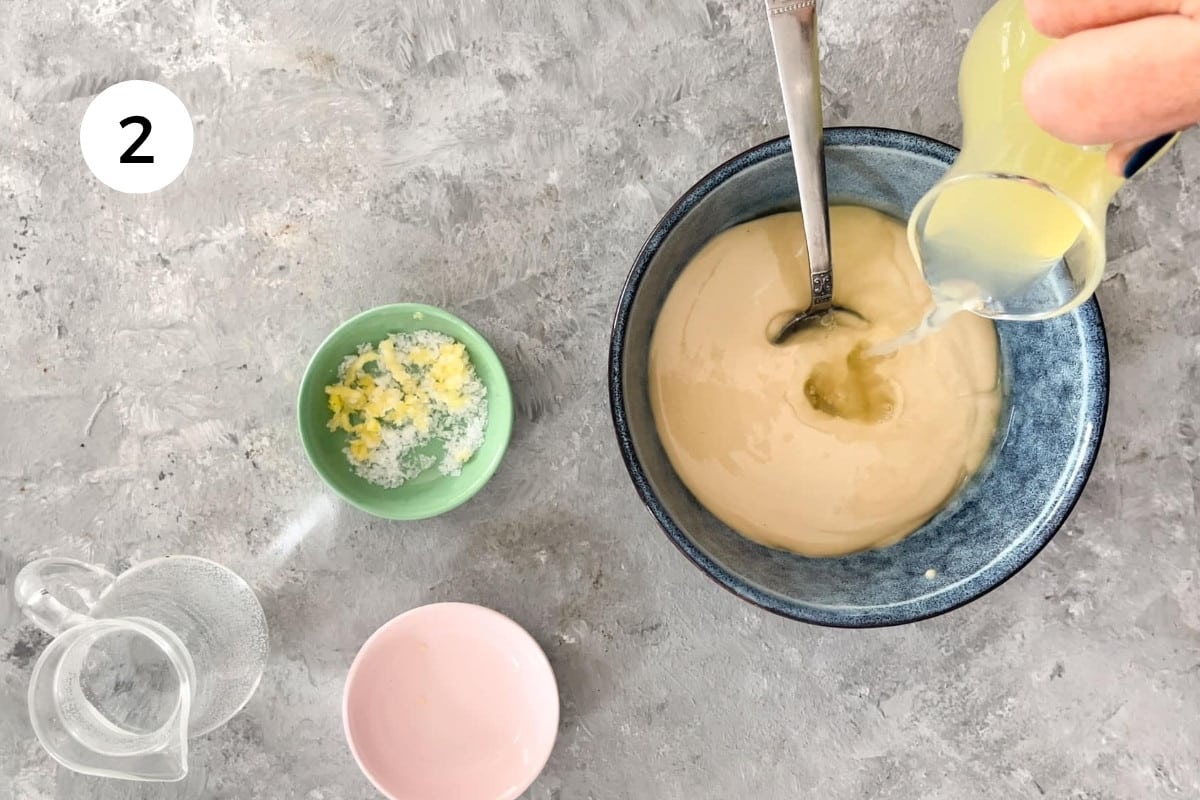A white hand is pouring lemon juice from a small glass jug into a bowl of tahini to make lemon tahini sauce.