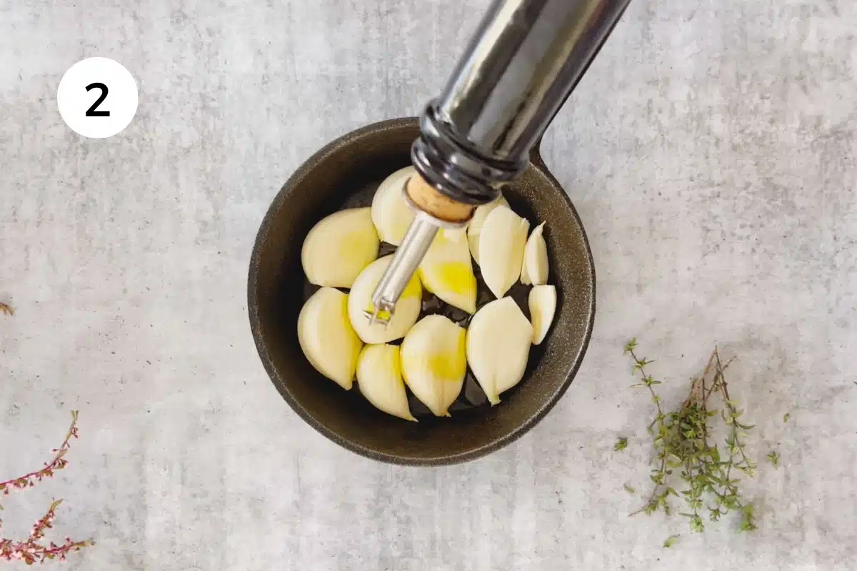 Olive oil is being poured from the bottle into a small cast iron frying pan filled with garlic cloves.
