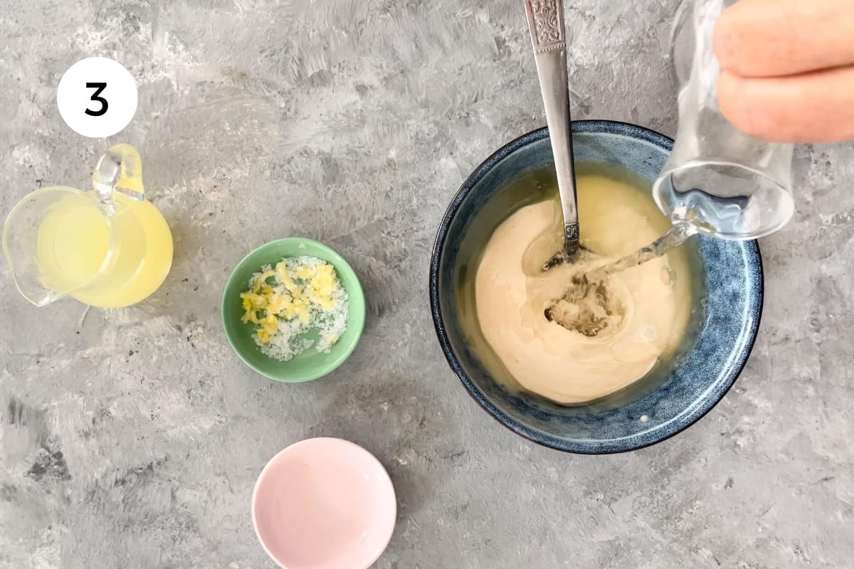 A white hand is pouring water from a small glass jug into a bowl of lemon tahini sauce to thin it out.