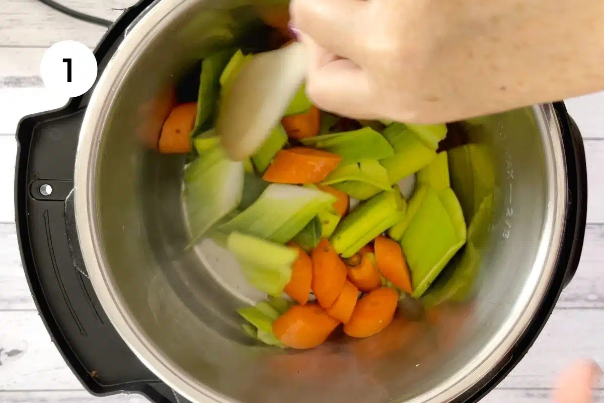 A white hand is stirring veg in the instant pot with a wooden spoon sautéing them for chicken soup.