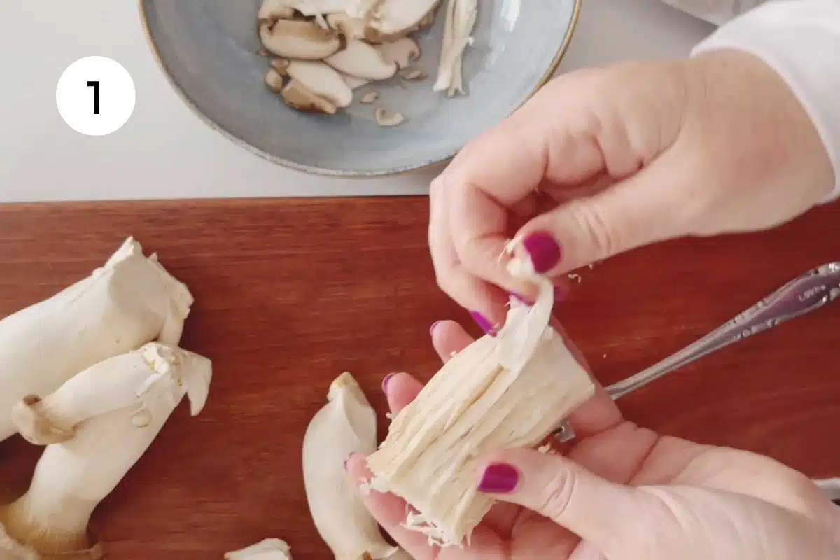 Two white hands are shredding the stem of a king oyster mushroom.