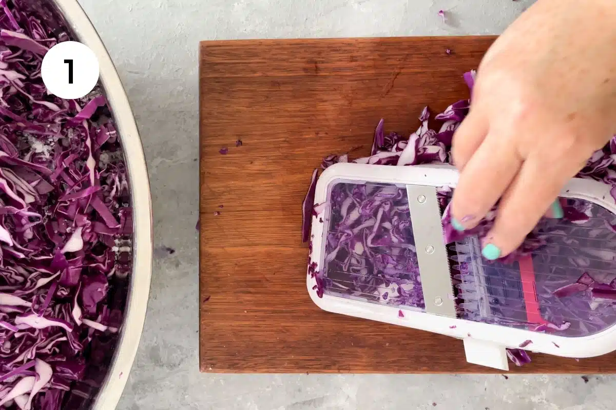 Two white hands are using a mandolin to slice up a red cabbage.