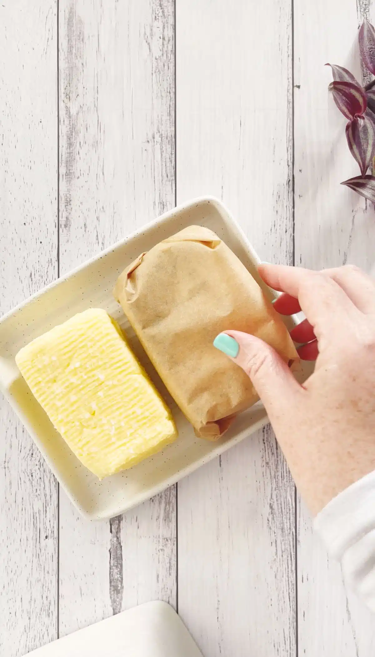 A white hand is placing a wrapped block of butter beside a block of yellow butter on a small butter dish.