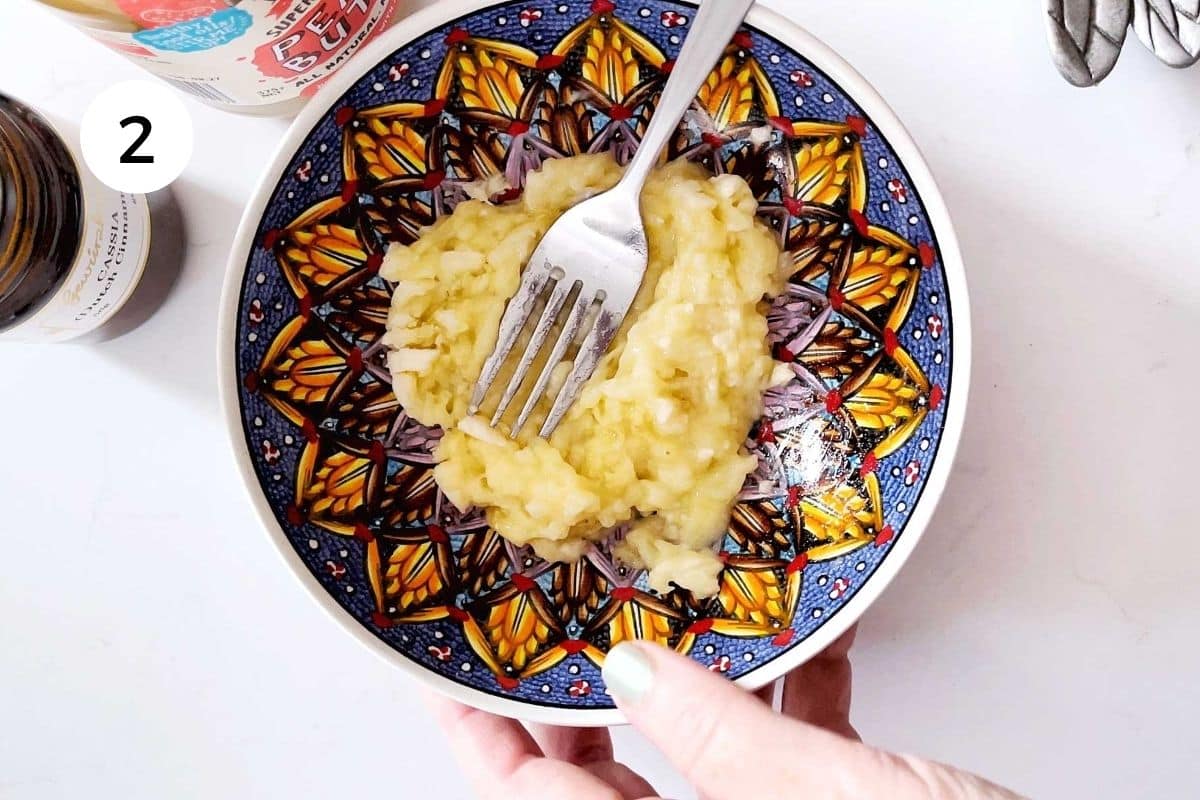 A white hand is holding a colourful bowl and mashing a banana in it with a silver fork.