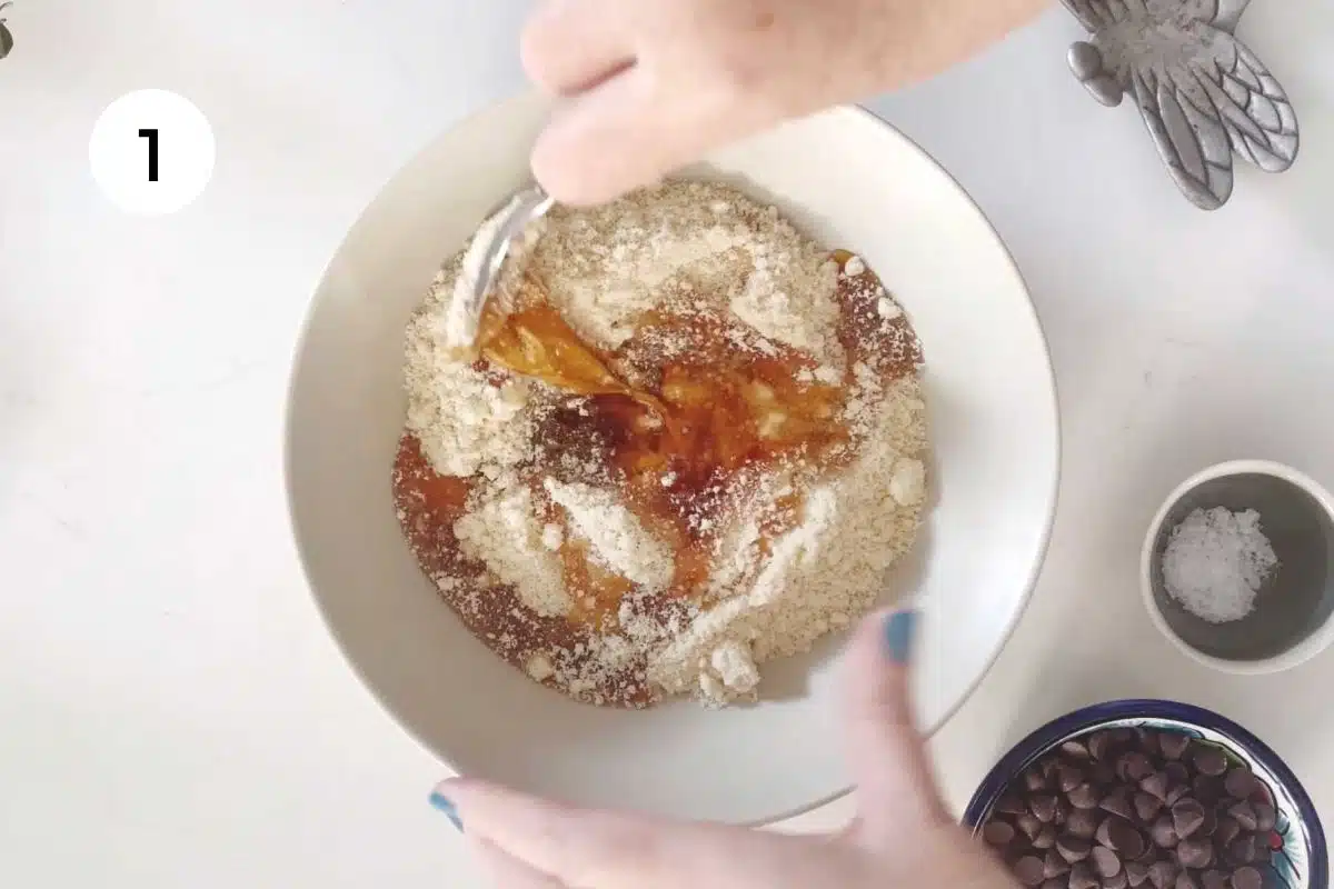 A white hand is using a silver spoon to mix a 4 ingredient peanut butter cookie batter in a large bowl.