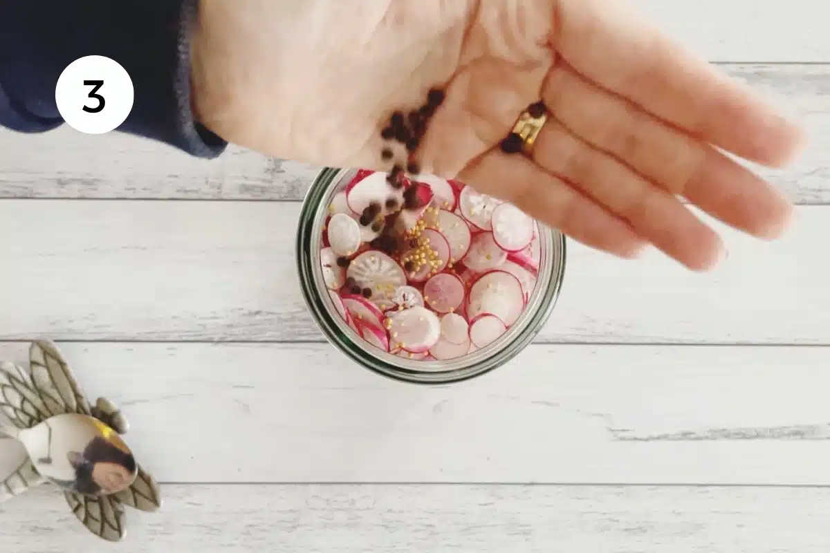 A white hand is dropping whole black peppercorns into pickled radish jar.