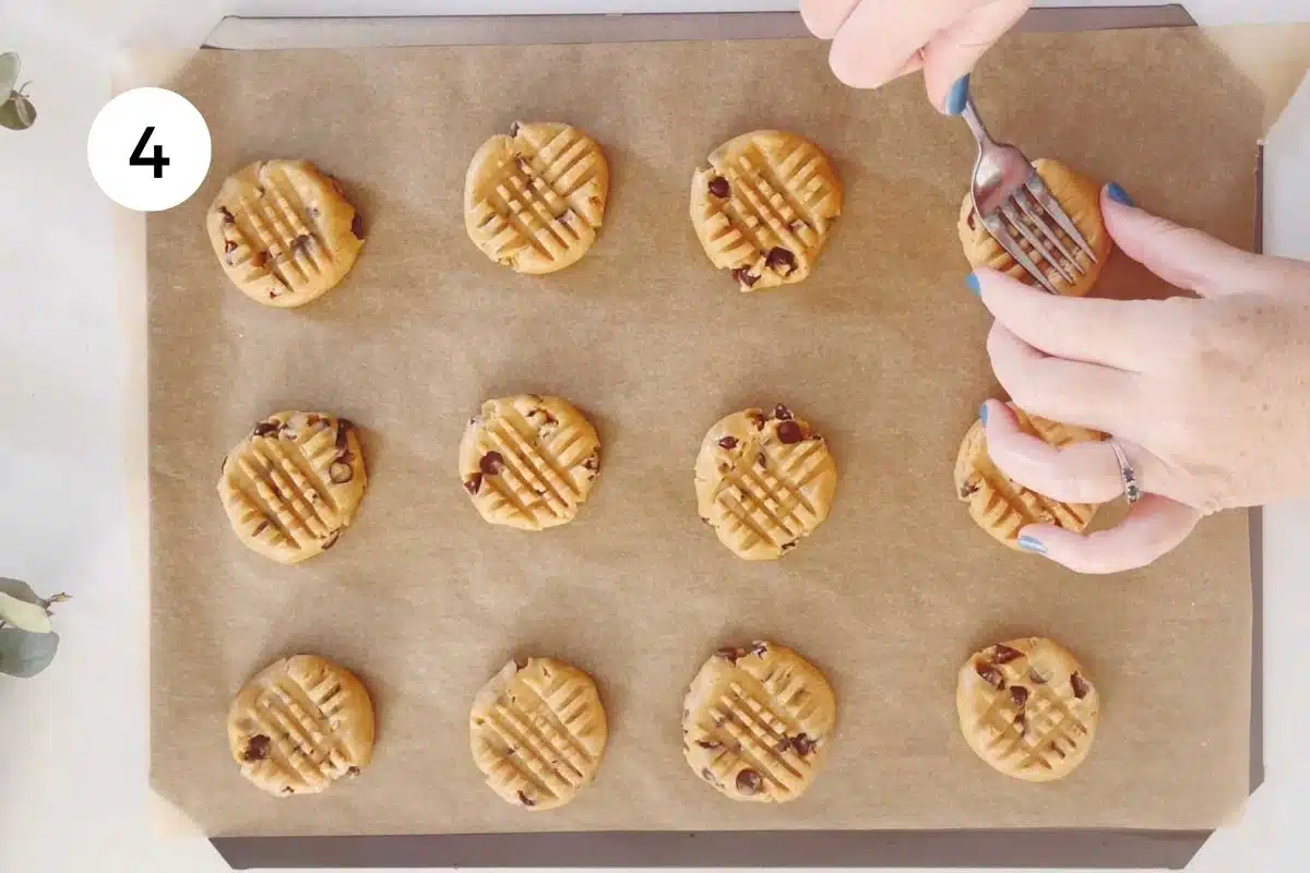 A white hand is using a silver fork to press down on cookie dough balls and flatten them out with a criss-cross pattern.