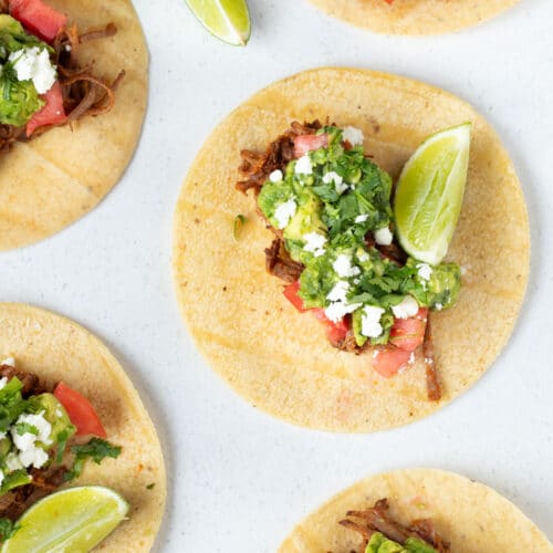 Corn tacos are topped with pulled brisket, tomato, guacamole, and feta cheese.