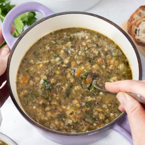 A white hand is stirring a small pot of barley and lentil soup with a silver spoon.