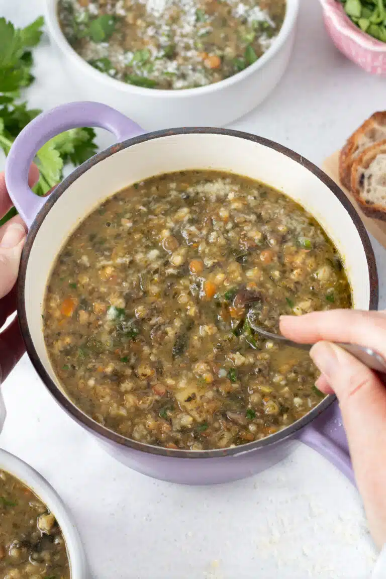 A white hand is stirring a small pot of barley and lentil soup with a silver spoon.