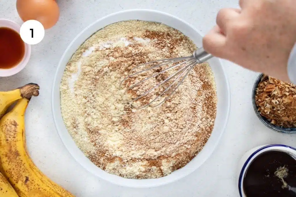 A white hand is using a whisk to stir together dry ingredients in a large white bowl for gluten free muffins.