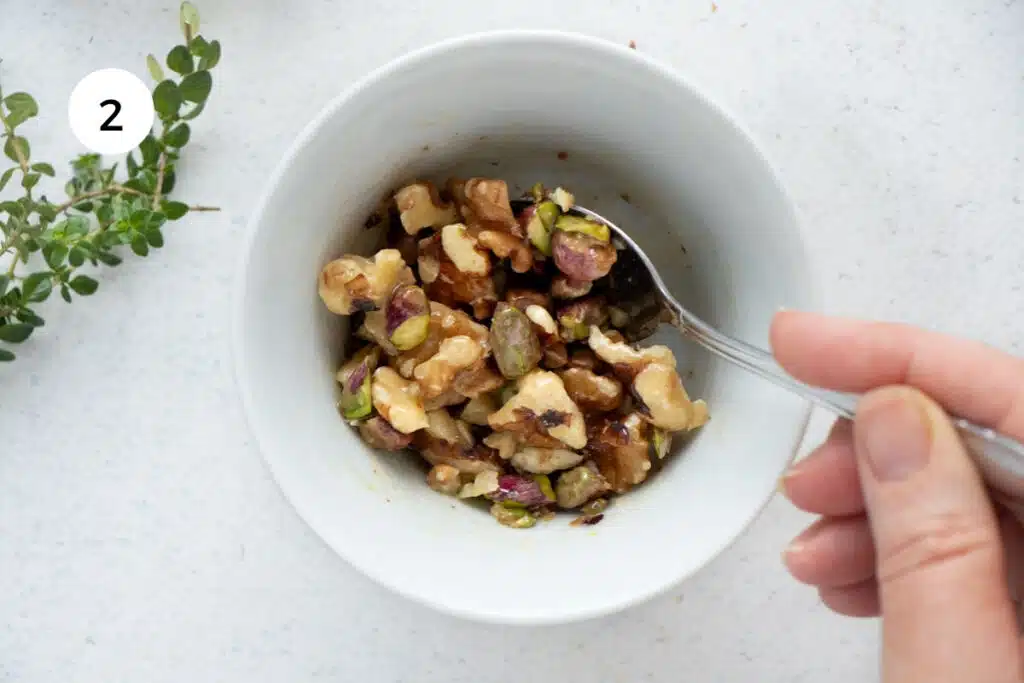 A white hand is using a spoon to stir together nuts, honey, and spices in a small bowl.
