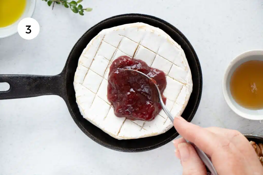 A white hand is using a teaspoon to dollop jam on top of a wheel of brie in a small cast iron skillet.