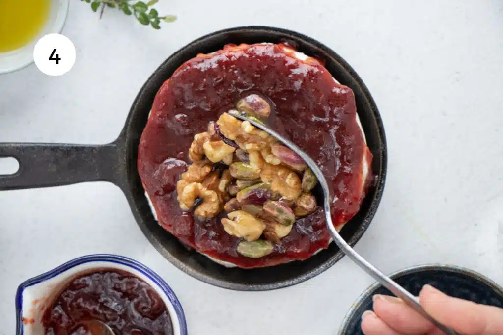 A white hand is spooning a spiced honey nut mix over the top of a wheel of brie covered in jam.