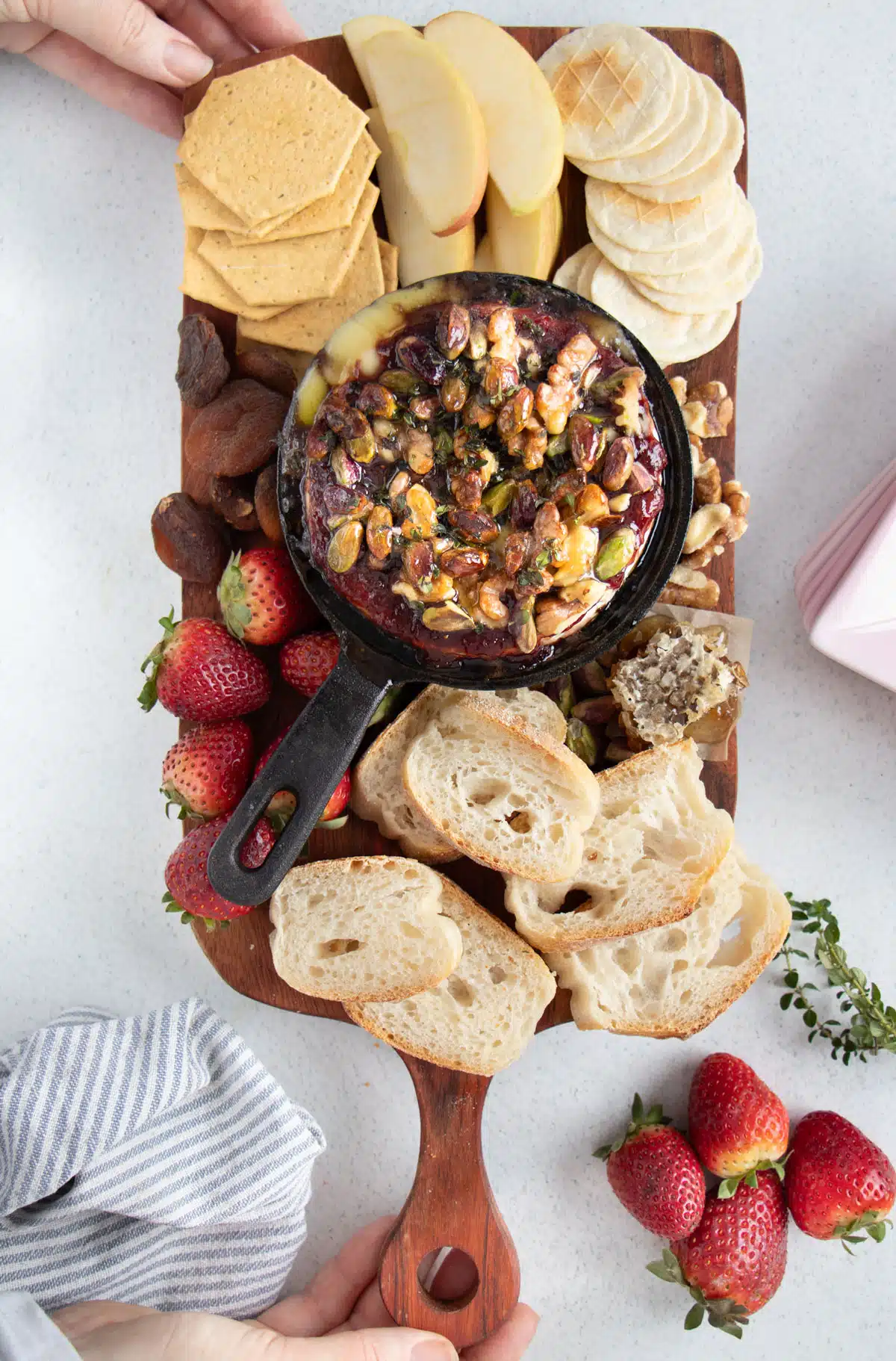 A snack board filled with baked brie with jam in a skillet, crackers, strawberries and sliced baguette and apple.