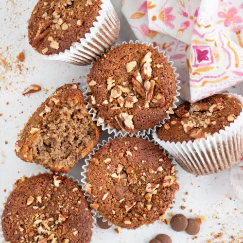 Several Almond Flour Banana Muffins are sitting on a table with brown sugar and nuts scattered around them.