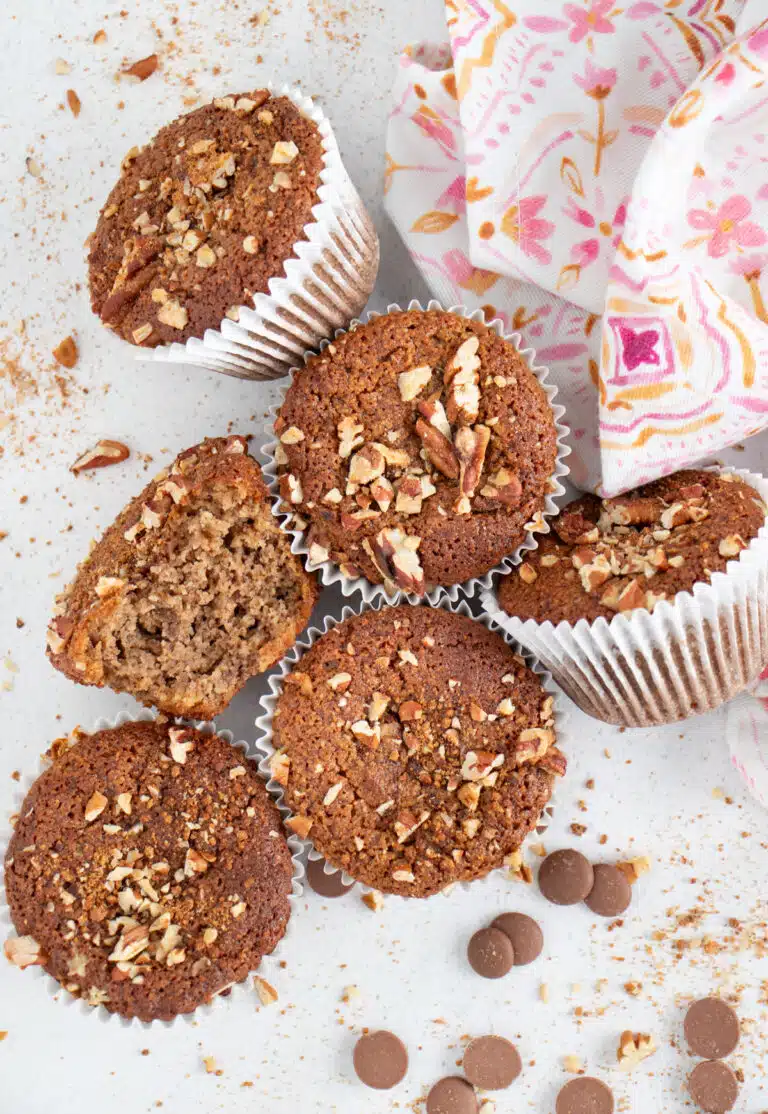 Several Almond Flour Banana Muffins are sitting on a table with brown sugar and nuts scattered around them.