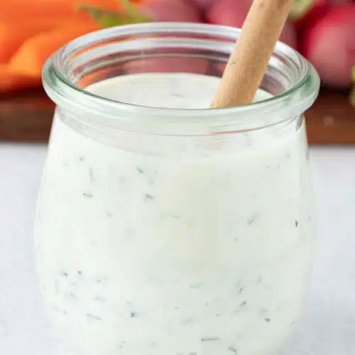 A small jar of Greek Yogurt Ranch Dressing is on a table with a wooden spoon in it.