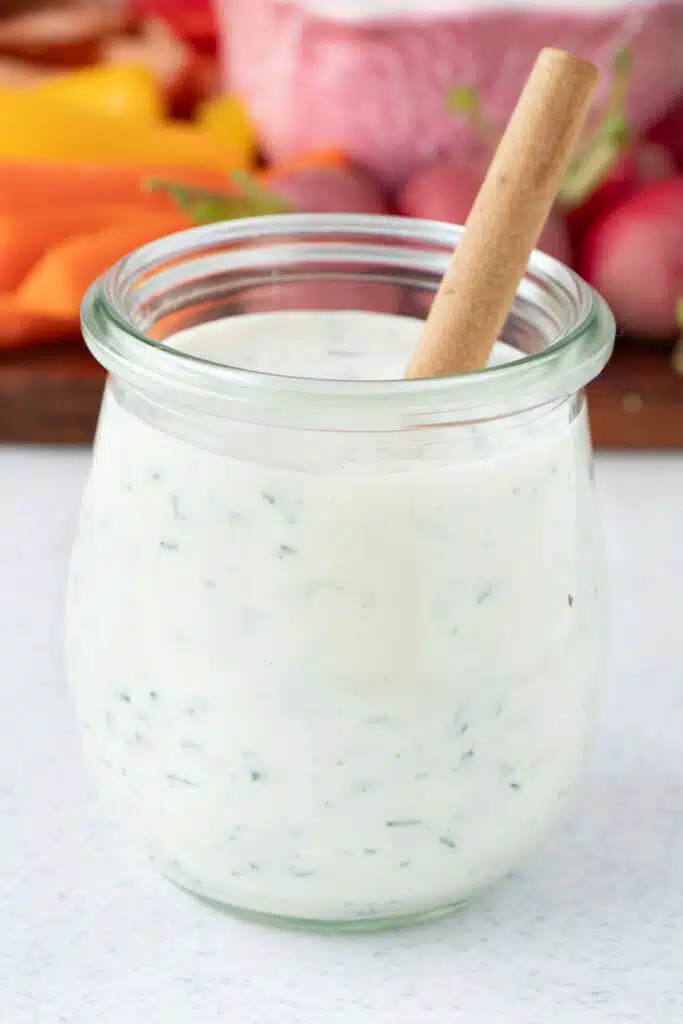 A small jar of Greek Yogurt Ranch Dressing is on a table with a wooden spoon in it.