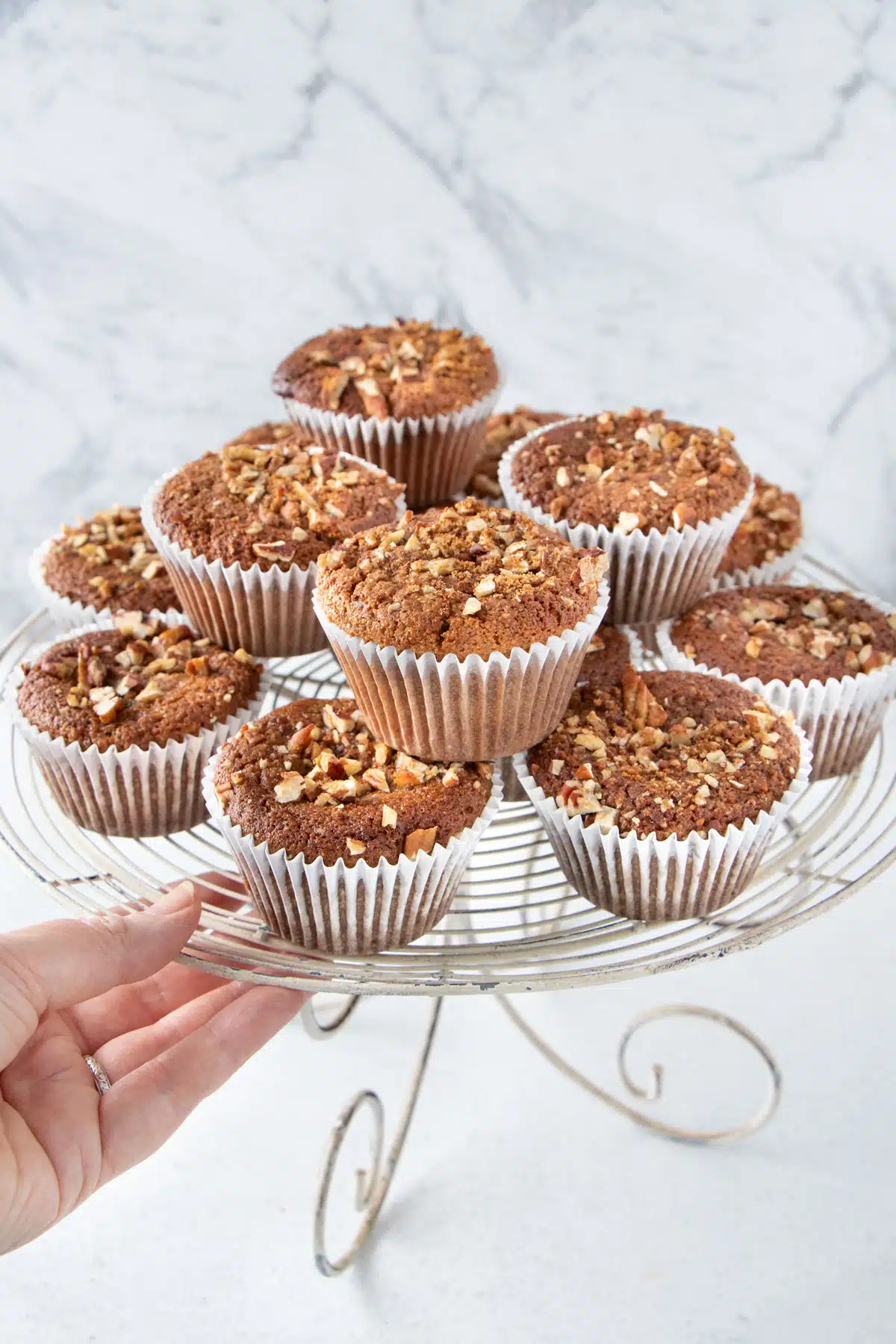 A white hand is placing a cake stand on a table piled high with almond flour banana muffins.