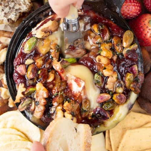 A white hand is dipping a small knife into a baked brie with jam to serve some on a slice of baguette.
