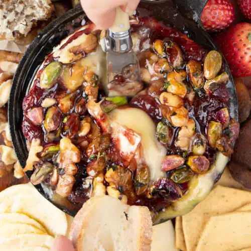 A white hand is dipping a small knife into a baked brie with jam to serve some on a slice of baguette.