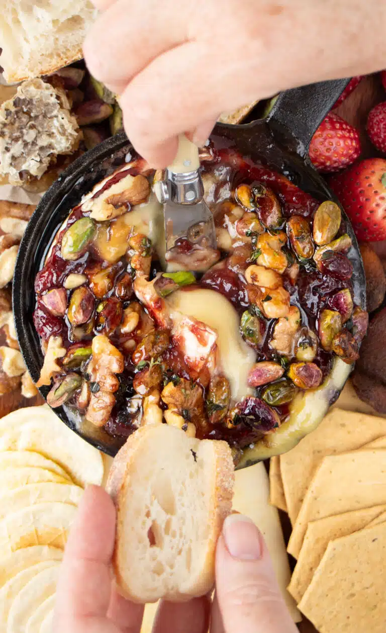 A white hand is dipping a small knife into a baked brie with jam to serve some on a slice of baguette.