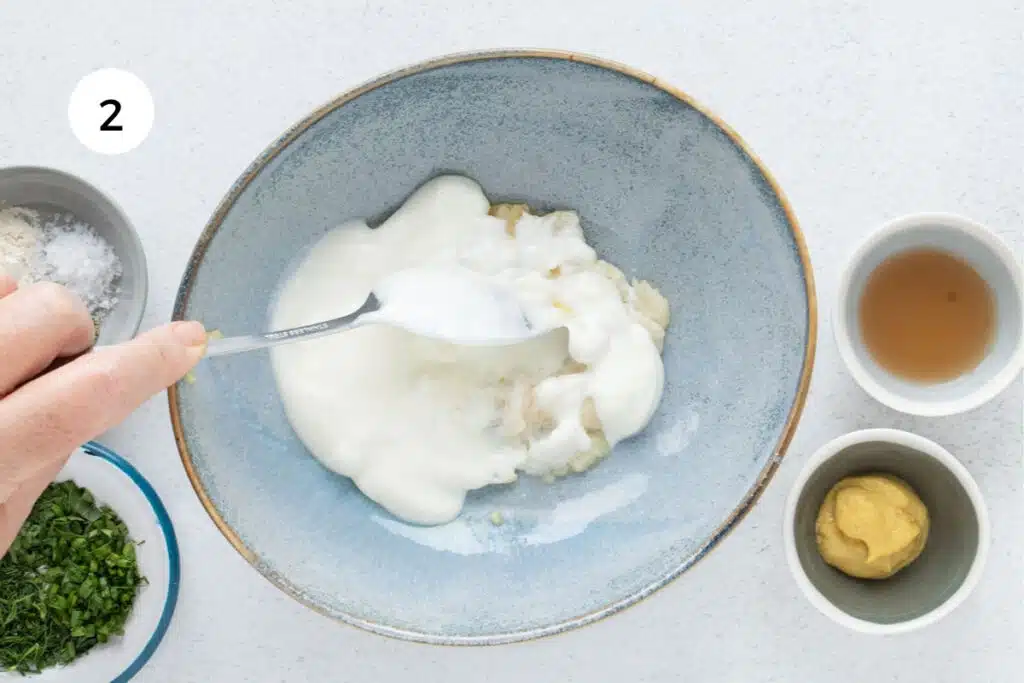 A white hand is using a silver spoon to stir together roasted garlic cloves and Greek yogurt.