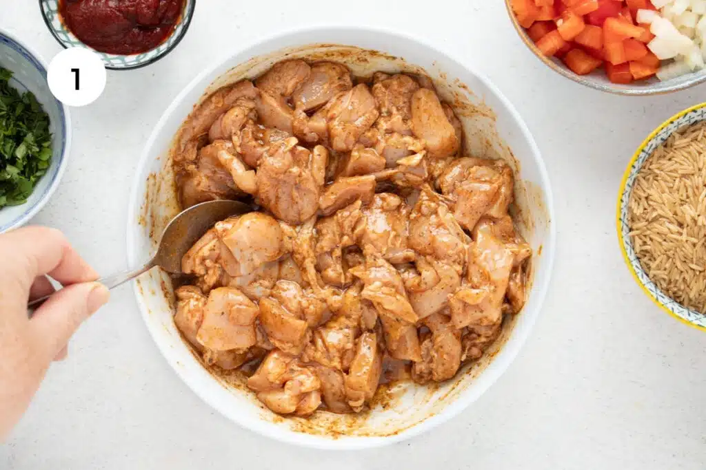 A white hand is using a silver spoon to toss chopped chicken thigh in marinade in a white bowl.