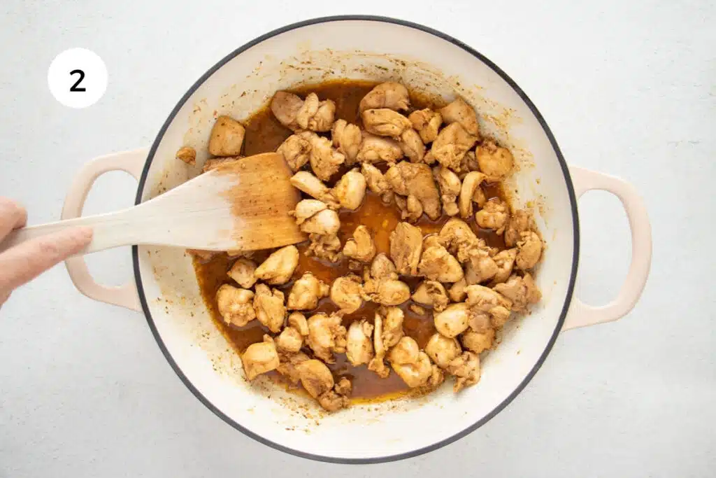 A white hand is using a wooden spoon to stir chicken pieces in a large French sauté pan.