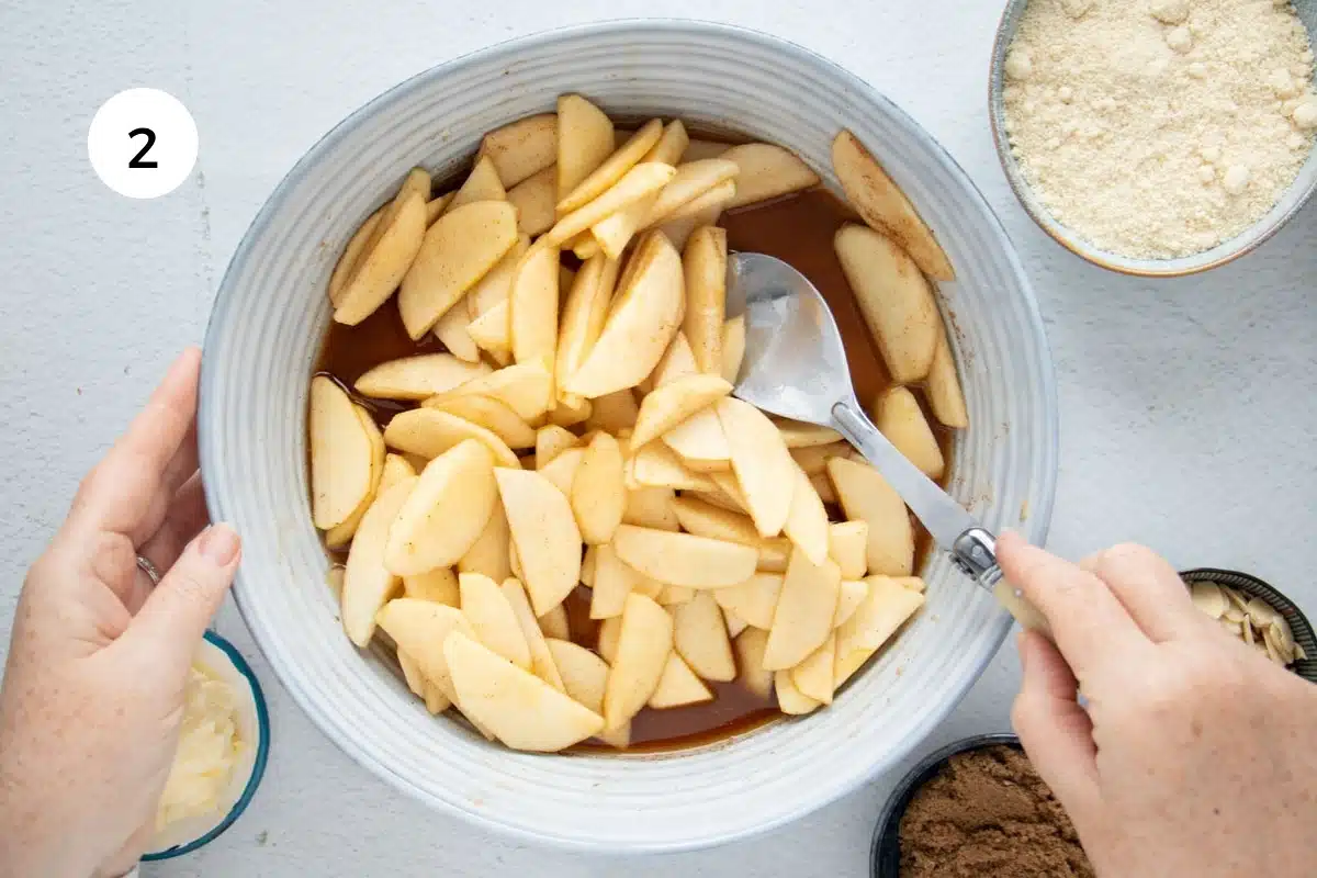 A white hand is stirring apple in spices and juice with a large spoon in a large blue grey bowl.