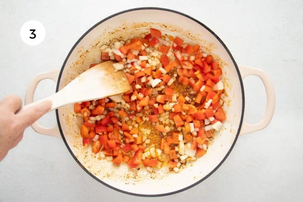 A white hand is using a wooden spoon to sauté chopped bell pepper and onion in a large pan.