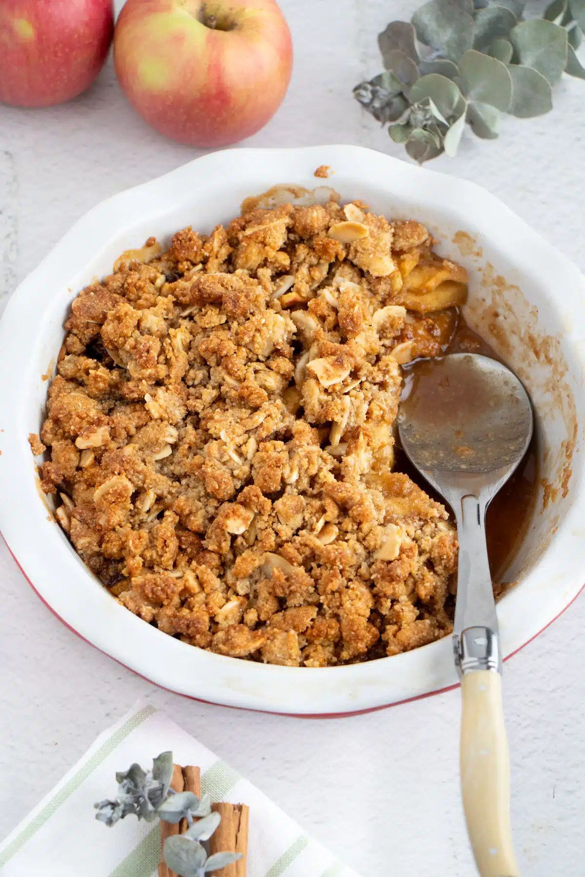 A pie dish full of apple crisp without oats with a big serving removed. A large spoon sits in the empty serving space.