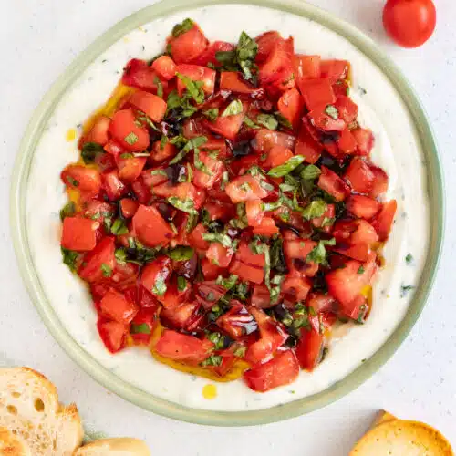 Bruschetta Dip is layered in a green bowl surrounded by sliced baguette and crackers.