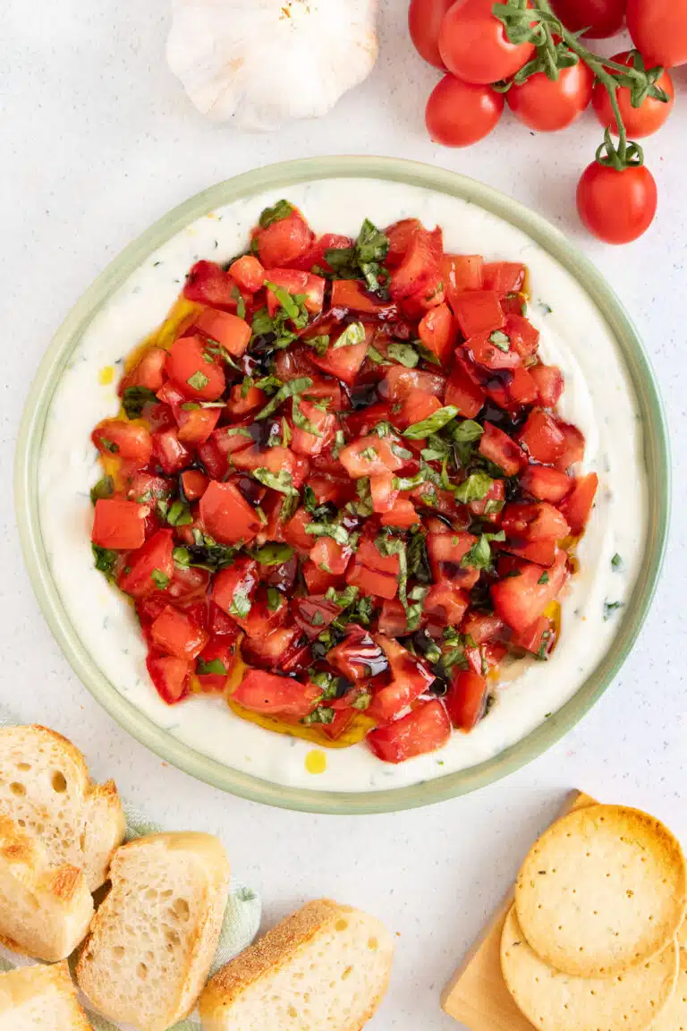 Bruschetta Dip is layered in a green bowl surrounded by sliced baguette and crackers.