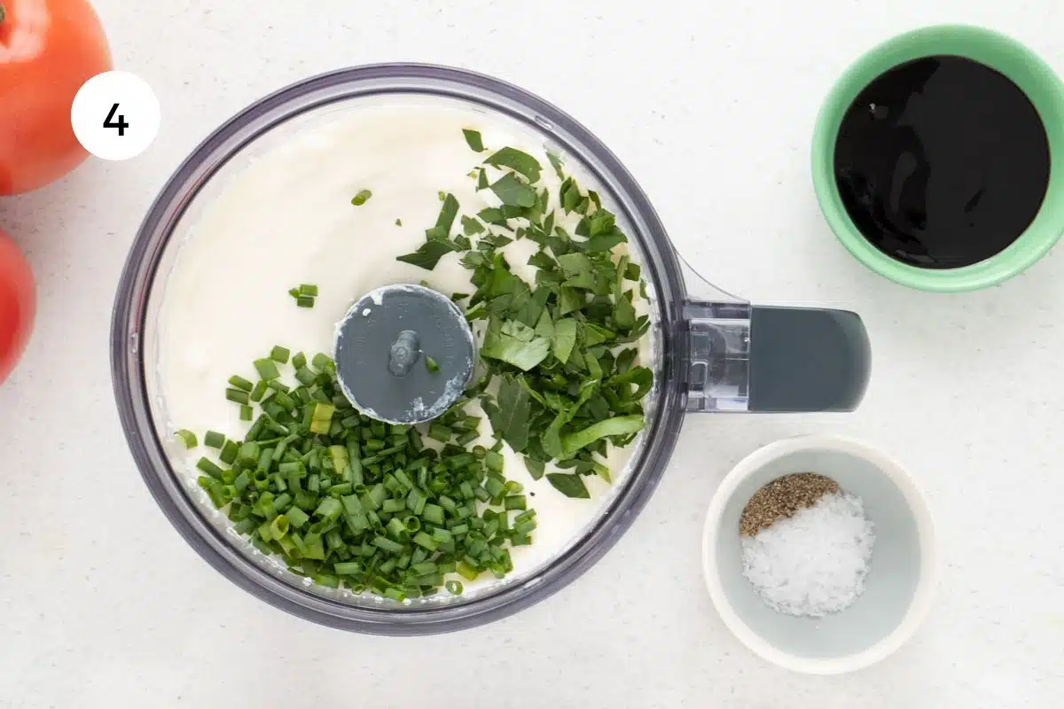 Whipped ricotta sits in the bowl of a small food processor topped with chopped fresh herbs.