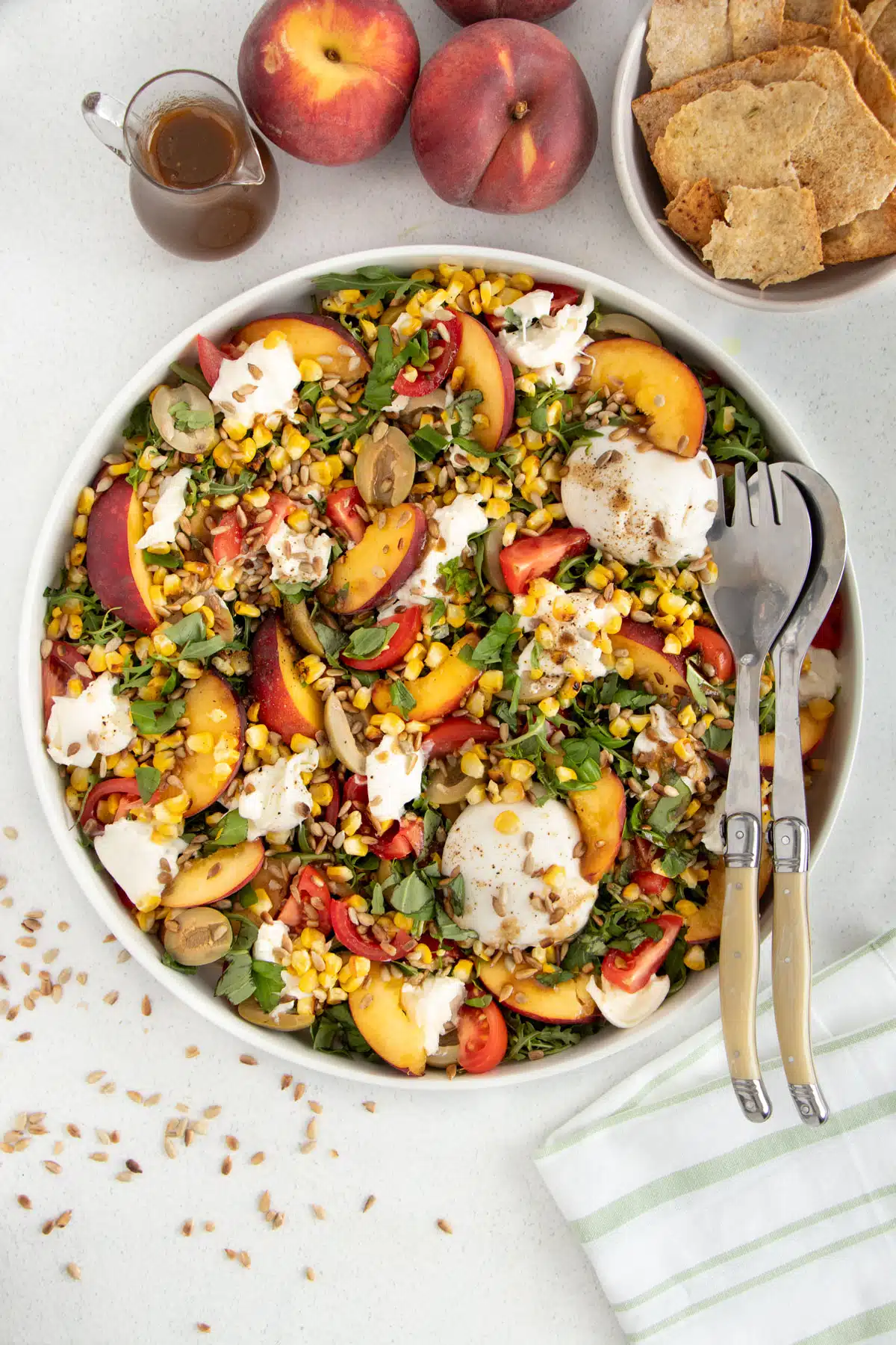 A large platter is filled with burrata peach salad, and two salad servers are sitting on the right side of the dish.