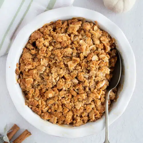 A round pie dish with a golden Apple Crisp Without Oats sits on a white table. A silver spoon is dug into the crisp.