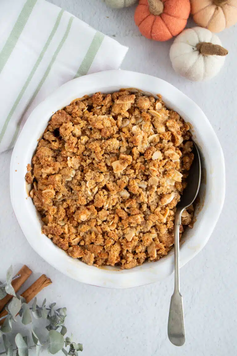 A round pie dish with a golden Apple Crisp Without Oats sits on a white table. A silver spoon is dug into the crisp.