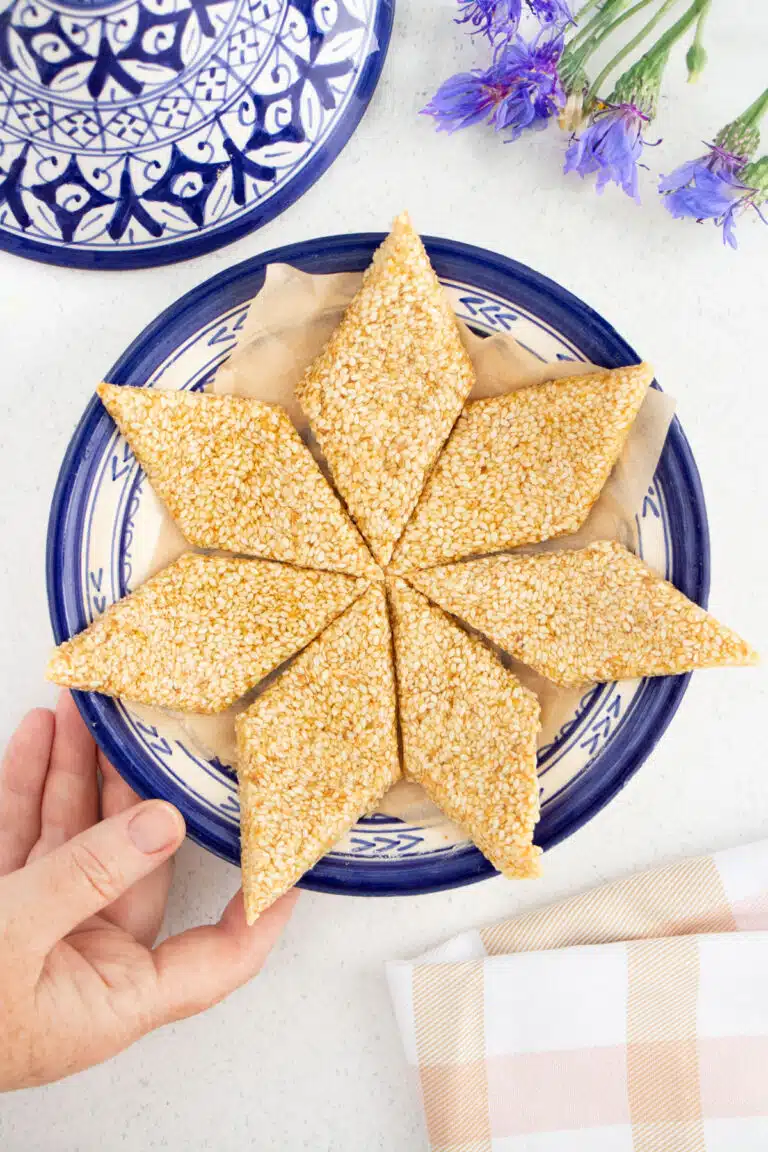 A white hand is placing a blue dish full of diamond shaped sesame candy onto a table.