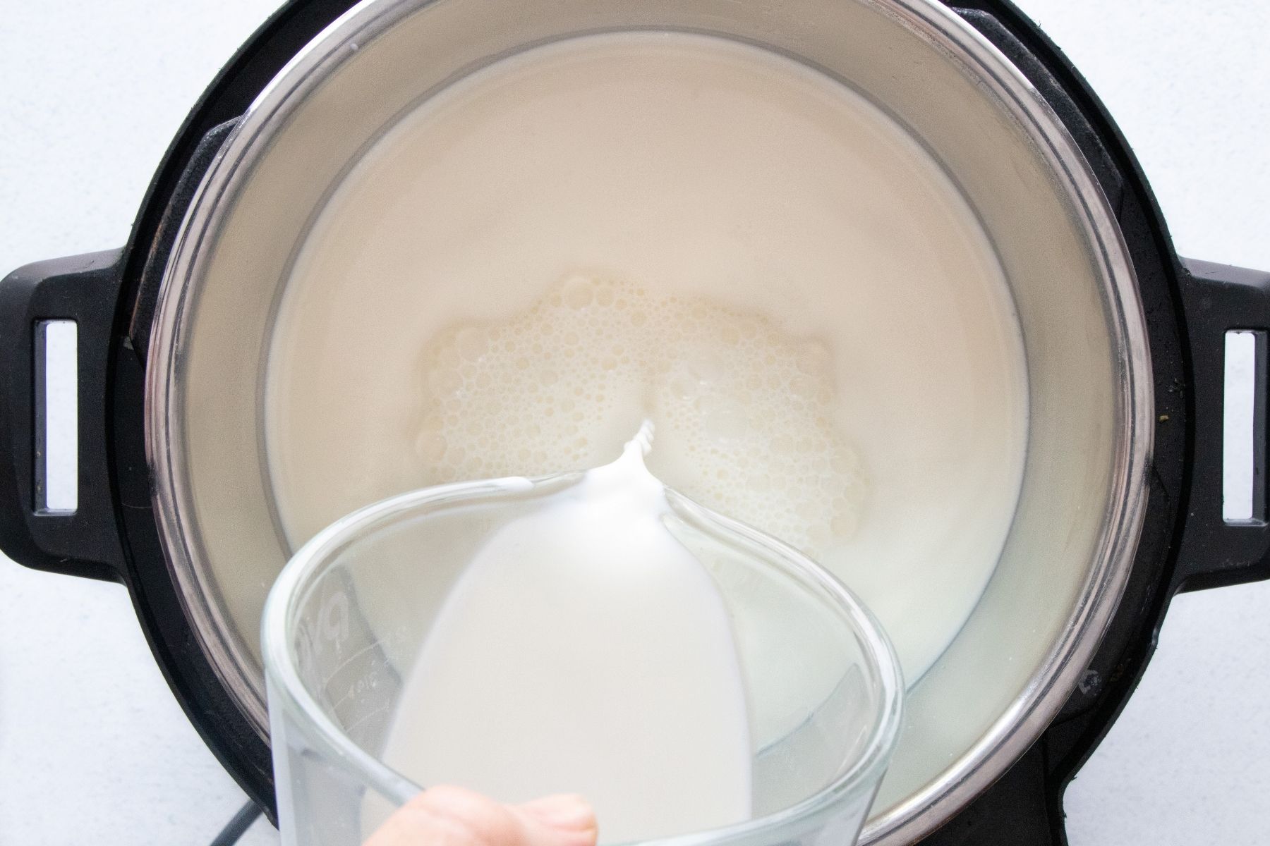 A white hand is pouring a jug of milk into the inner pot of an instant pot.
