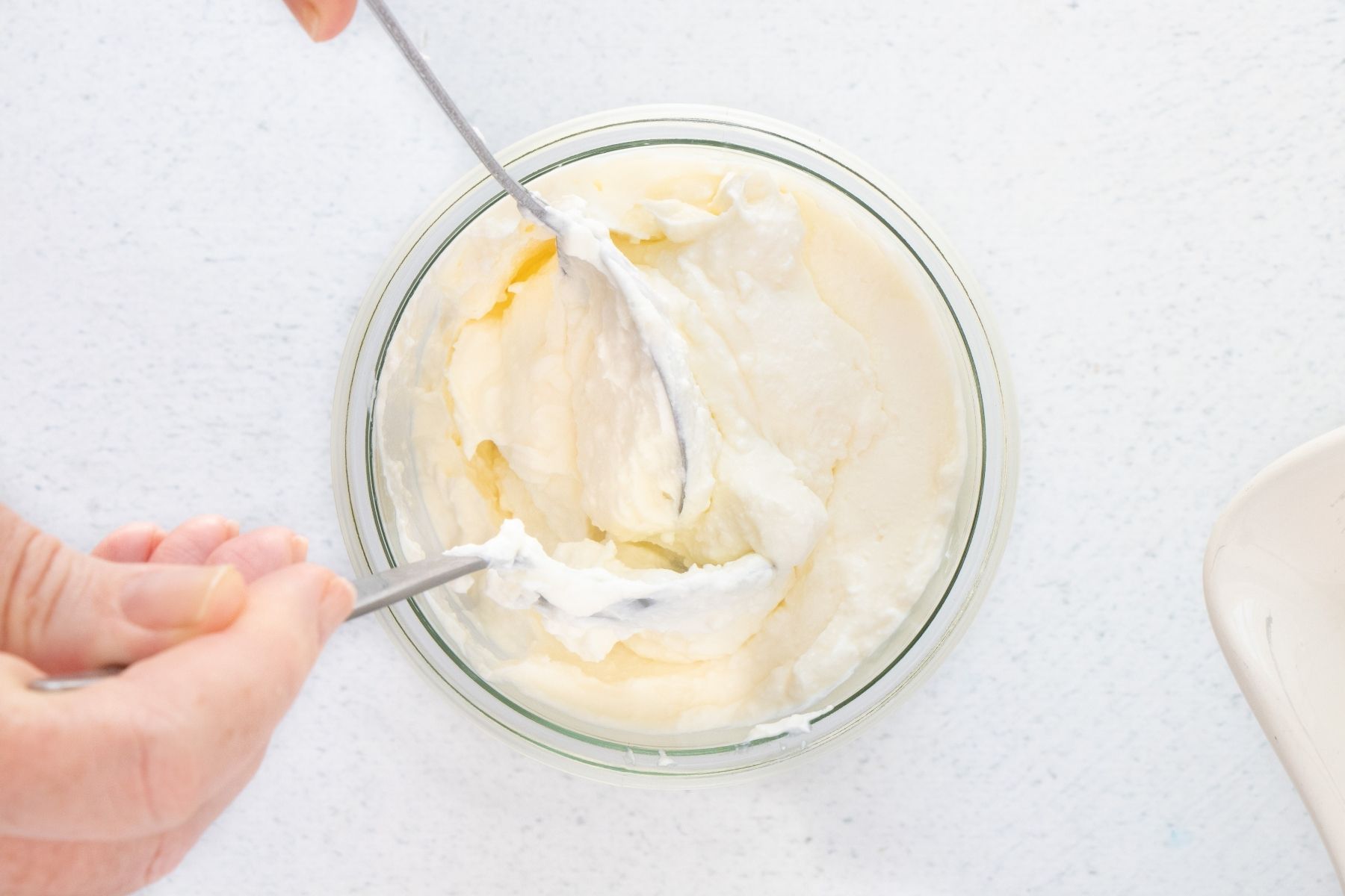 Two white hands are using silver spoons to spoon thickened Greek yoghurt into a glass jar.
