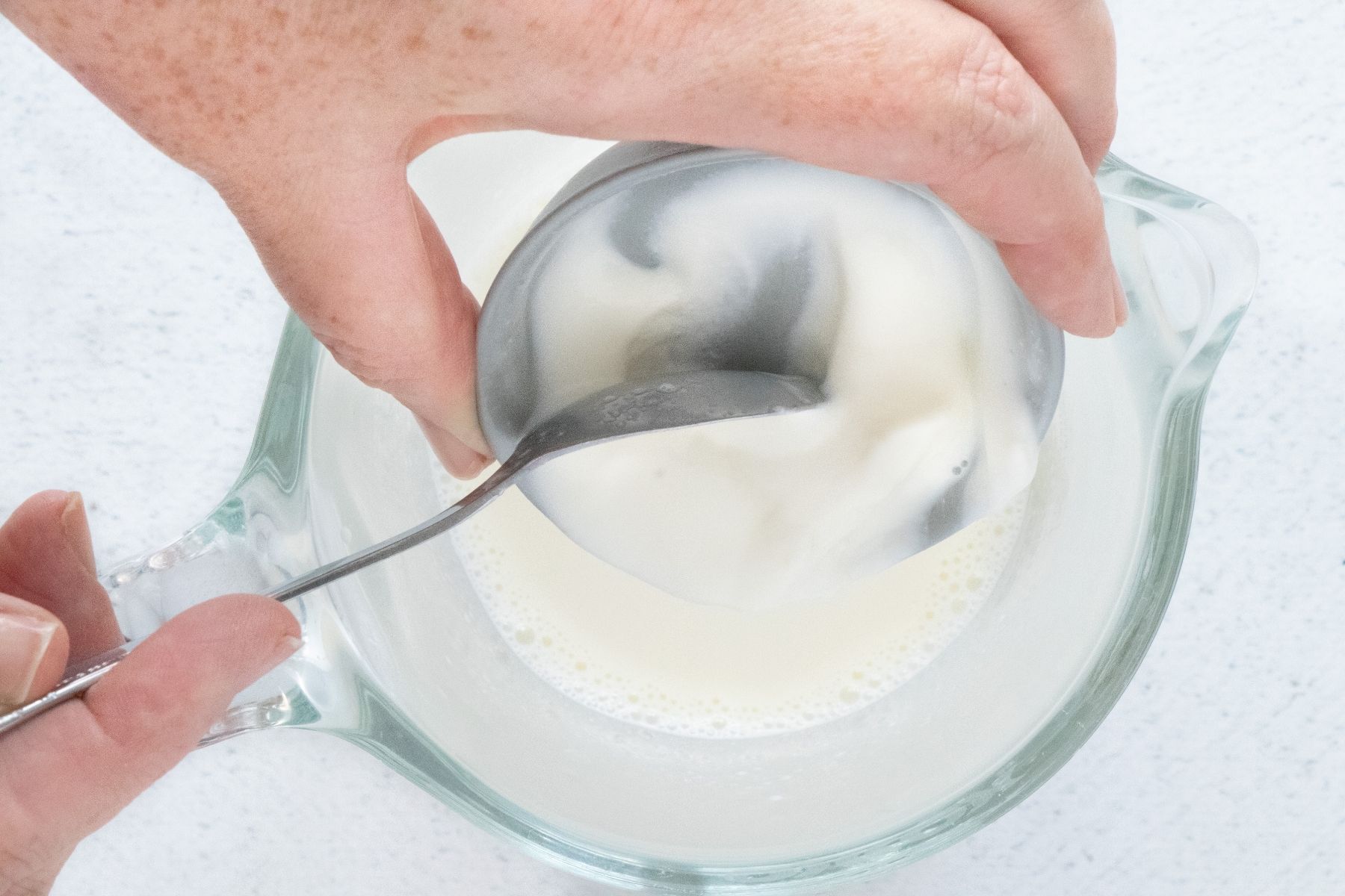 A white hand is tipping a small amount of yogurt into warmed milk in a jug to make a yogurt starter.