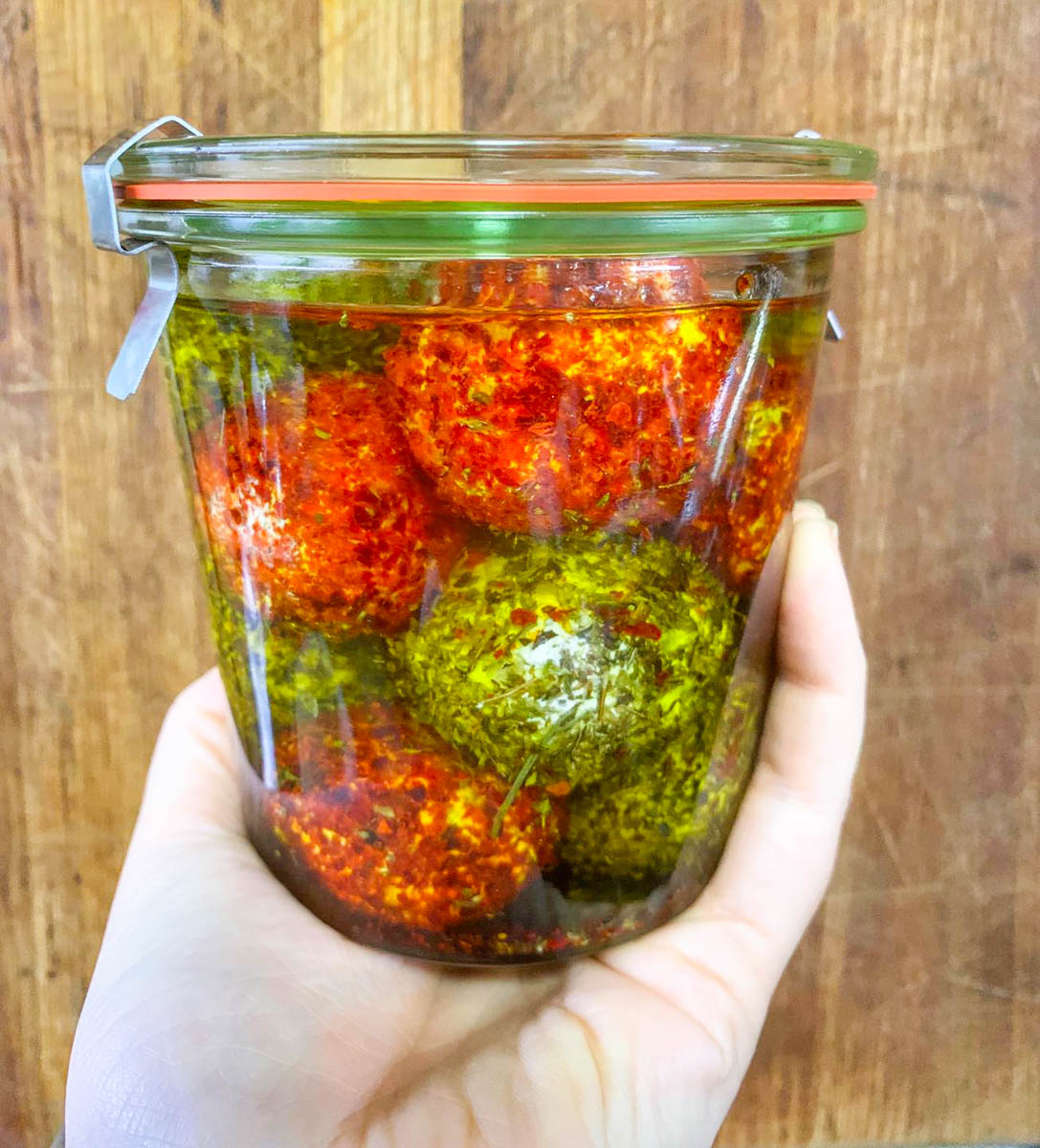 A white hand is holding up a jar filled with labneh balls rolled in spices and covered in olive oil.
