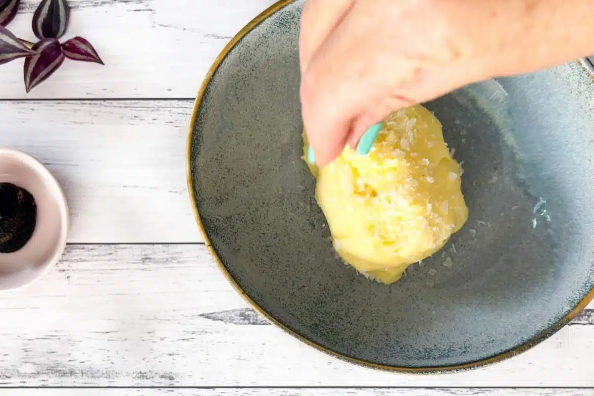 A blue bowl full of butter is sitting in the middle of a white wooden table. A white hand is sprinkling salt over the top.