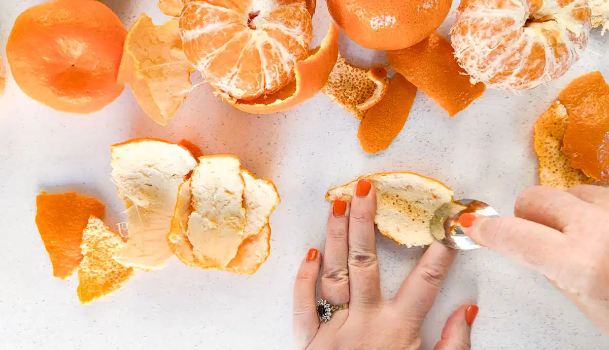 A white hand is holding a piece of mandarin peel while the other hand scrapes out the pith with a teaspoon.
